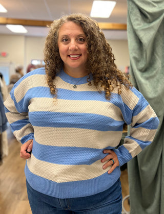 A woman with curly brown hair wearing a blue and cream striped knit sweater and blue jeans, standing in a fitting room or boutique.