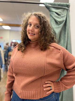 A woman with curly brown hair wearing a dusty rose ribbed knit mock neck sweater and blue jeans, standing in what appears to be a fitting room or boutique.