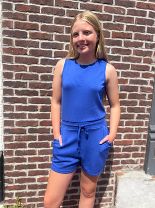 A young girl with blonde hair stands in front of a red brick wall wearing a bright blue sleeveless top and matching blue shorts.