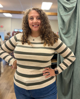 A woman with curly brown hair wearing a beige and dark olive green striped long-sleeve shirt and blue jeans, standing in a fitting room with her hands on her hips.
