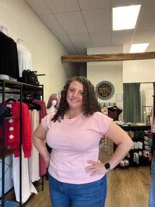 A woman with curly brown hair wearing a light pink short-sleeve top and blue jeans, smiling while posing in a retail store environment.