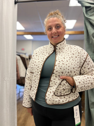 A woman wearing a cream quilted jacket with a brown floral pattern and a teal top, standing in a clothing store.