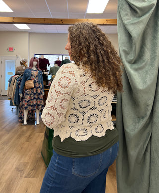 A woman with curly brown hair is shown from behind wearing a cream crochet lace top and blue jeans inside a clothing store. A sage green curtain is visible on the right side of the frame.