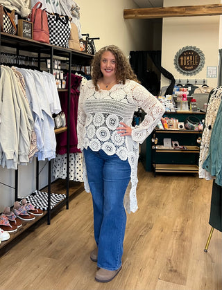 A woman with curly brown hair stands in a boutique wearing a white crochet long-sleeve top with a scalloped hem and blue jeans. The shop has wooden floors, clothing racks, and shelves in the background.