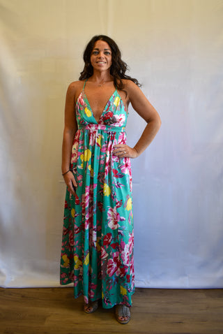 A woman modeling a vibrant, multicolor floral maxi dress with a deep V-neckline and thin spaghetti straps, standing against a white fabric backdrop.