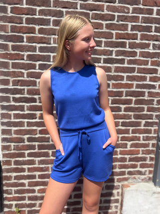 A young woman wearing a bright royal blue sleeveless top and matching shorts with a drawstring waist, standing against a red-brown brick wall.
