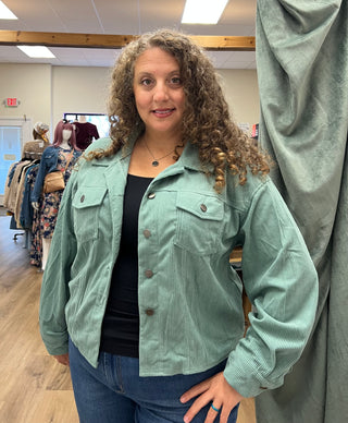 A woman with curly brown hair wearing a sage green corduroy button-up jacket over a black top and blue jeans, standing inside a clothing store.