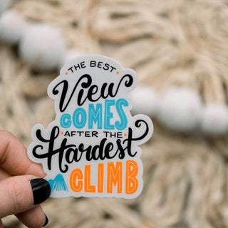 A hand with black nail polish holds a die-cut sticker that reads 'THE BEST View COMES AFTER THE Hardest CLIMB' in black, blue, and orange lettering, with a blurred rope background.