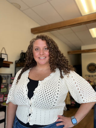A smiling woman with curly brown hair wearing a white crochet cardigan over a black top and blue jeans, standing in a retail store interior.