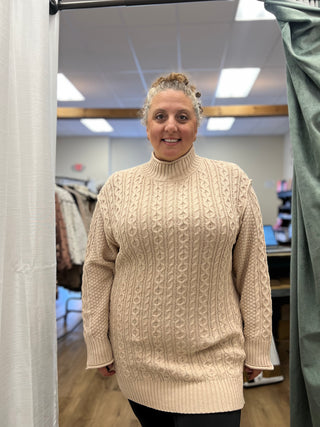 A woman wearing a cream cable-knit turtleneck sweater dress stands in a fitting room or boutique interior, with clothing racks and store fixtures visible in the background.