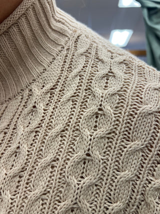Close-up of a cream-colored cable-knit sweater showing the detailed knit pattern and ribbed collar.