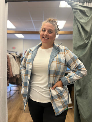 A woman posing in a fitting room wearing a light blue and white plaid button-up shirt over a white t-shirt and black pants.
