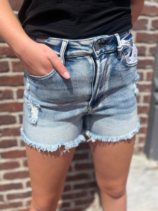 Light-wash distressed denim shorts with a frayed hem and ripped details, shown on a model standing against a brick wall.
