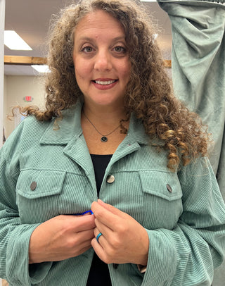A smiling woman with curly brown hair wearing a sage green corduroy jacket over a black top, standing indoors.