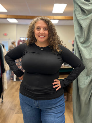 A woman with curly brown hair wearing a fitted black long-sleeve top and blue jeans, standing in a clothing store with her hands on her hips.