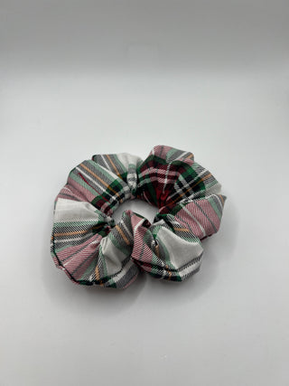 A fabric hair scrunchie featuring a classic tartan plaid pattern in green, red, pink, and white, shown on a neutral light gray background.