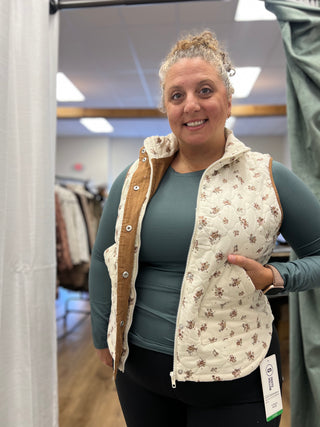A woman wearing a cream quilted vest with a brown floral pattern and tan buttoned lining, layered over a sage green long-sleeve shirt and black pants, standing in a retail fitting room.