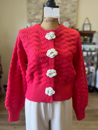 A coral pink cable-knit cardigan sweater with four white crocheted flower buttons, displayed on a cream dress form mannequin in a softly blurred indoor setting.