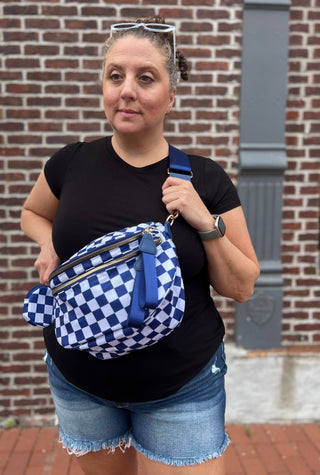 A woman wearing a black t-shirt and denim shorts stands in front of a brick wall, holding a blue and white checkered crossbody bag with a blue strap.