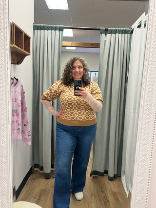 A woman takes a mirror selfie in a fitting room wearing a beige and brown geometric patterned knit sweater with light pink long sleeves, paired with blue jeans and white sneakers.