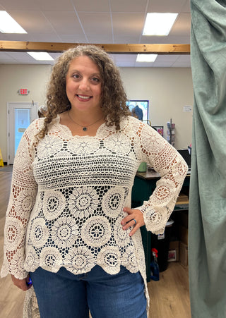 A woman with curly brown hair wearing a cream-colored crocheted long-sleeve top and blue jeans, standing in an indoor room with office desks and fluorescent lights in the background.