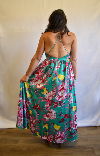 A woman viewed from behind wearing a sleeveless maxi dress with a bright floral print on a teal-green background. The dress has thin halter-style straps and an open back, and she is holding up the skirt to show its full flowy shape.