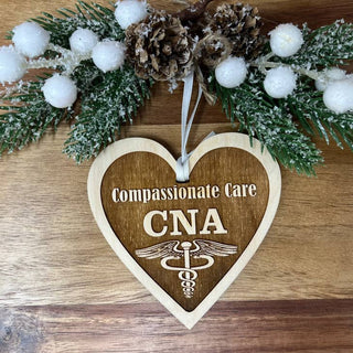 A heart-shaped wooden ornament engraved with 'Compassionate Care CNA' and a caduceus symbol, hanging from a white ribbon. The ornament is displayed on a wooden surface decorated with a festive wreath featuring green pine branches, white faux snow, and pinecones.