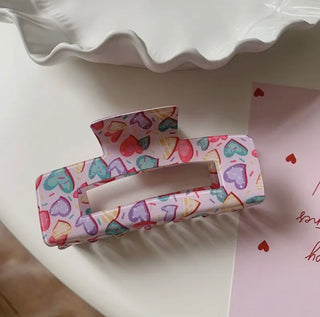 A pastel-colored hair claw clip with a colorful heart pattern, resting on a white surface next to a pink card with red hearts.