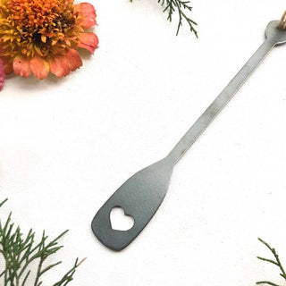 A long-handled metal tool with a paddle-shaped head and a heart-shaped cutout, photographed on a white textured surface with decorative flowers and greenery in the corners.