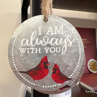A round hanging ornament with a rustic silver-gray distressed finish, featuring the phrase 'I AM always WITH YOU' in white lettering and two red cardinal birds near the bottom. The ornament is suspended by a jute rope and photographed indoors against a home shelf background.