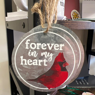 A round wooden hanging ornament with a rustic gray finish, featuring the words 'forever in my heart' in white script and a red cardinal bird silhouette. The ornament hangs from a natural jute rope in a home setting.