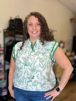 A woman with curly brown hair wearing a white sleeveless blouse with a green botanical toile pattern and green ruffled trim, paired with blue jeans, standing in a home setting.