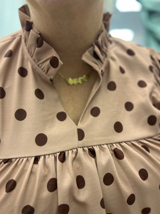 Close-up of a person wearing a beige blouse with dark brown polka dots and a ruffled high neckline, paired with a small yellow necklace.