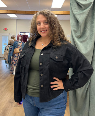 A woman with curly brown hair stands in a fitting room wearing a black corduroy jacket over an olive green top and blue jeans.