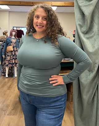 A woman with curly brown hair wearing a sage green long-sleeve ribbed top and blue jeans, standing inside a clothing store.