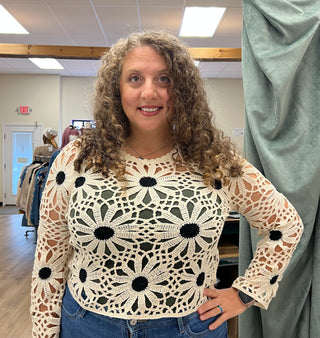A woman with curly brown hair wearing a cream crochet long-sleeve top with black floral motifs and blue jeans, standing in a fitting room.