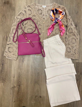 A styled flat-lay outfit on a wood floor featuring a beige crochet cardigan, cream-colored pants, a bright magenta crossbody bag, and a colorful orange, pink, and blue patterned silk scarf.