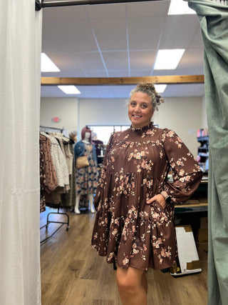 A woman modeling a brown long-sleeve mini dress with a pink and white floral pattern, standing in a fitting room.