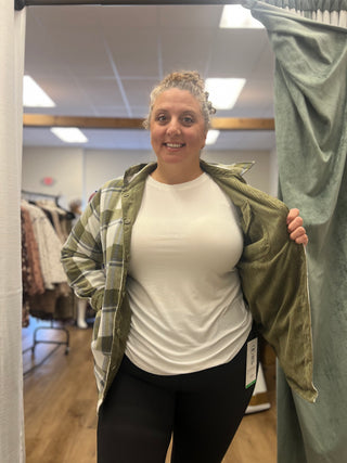 A woman in a fitting room wearing a white t-shirt, black leggings, and an olive green corduroy overshirt over a green plaid flannel shirt. She is holding the green curtain open on the right side of the frame.