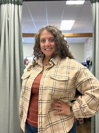 A woman with curly brown hair stands in a fitting room wearing a beige and brown plaid button-up shirt over a rust-colored top.