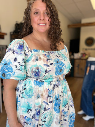 A woman wearing a white off-the-shoulder dress with a blue and teal floral print, standing in a home-like setting.