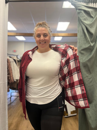 A woman in a fitting room wearing a white t-shirt and black leggings, holding up a red and white plaid flannel shirt over her shoulders. Clothing racks and a green curtain are visible in the background.