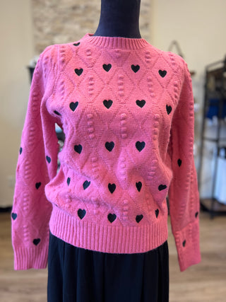A pink cable-knit sweater with black heart motifs, displayed on a black dress form mannequin in an indoor setting.