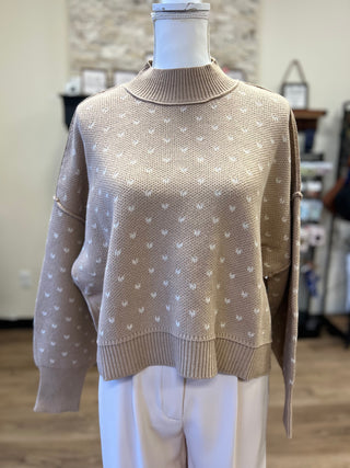 A beige knit sweater with a white heart pattern, displayed on a mannequin torso in an indoor boutique setting.