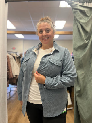 A woman wearing a light blue corduroy button-up shirt over a white t-shirt and black pants, standing in a fitting room.