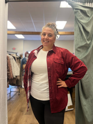A woman modeling a red corduroy button-up shirt over a white t-shirt and black leggings, standing in a fitting room with a green curtain.