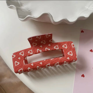 A rectangular hair claw clip with a coral-red and white heart pattern, resting on a white surface next to a white wavy dish.