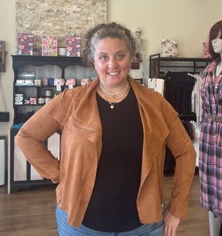 A woman wearing a brown suede moto jacket over a black top and blue jeans, standing in a boutique store.