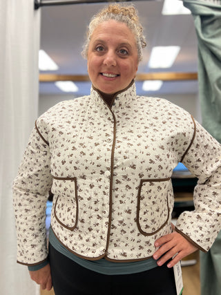 A woman wearing a cream-colored quilted jacket with a brown floral pattern and brown trim, standing in an indoor setting.