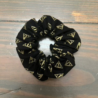 A black fabric scrunchie with a repeating gold Deathly Hallows triangle pattern, displayed on a dark wooden surface.